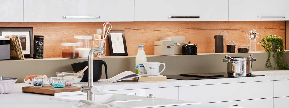 Zeitgemäße Küche mit weißen Fronten, Holz-Hintergrund, Arbeitsfläche mit Kochtöpfen, Geschirr und Küchenutensilien, freundliche Beleuchtung.